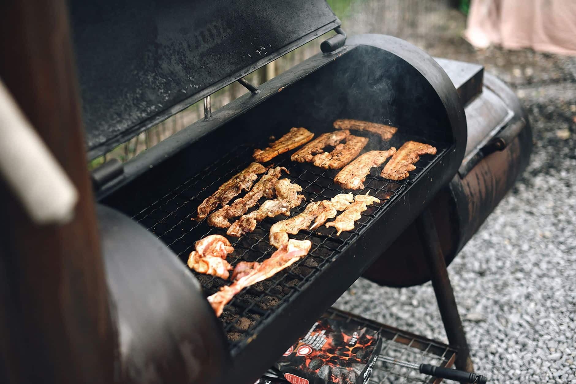 Close-up of a barbecue grill outdoors with various meat strips cooking, exuding rich smoke.