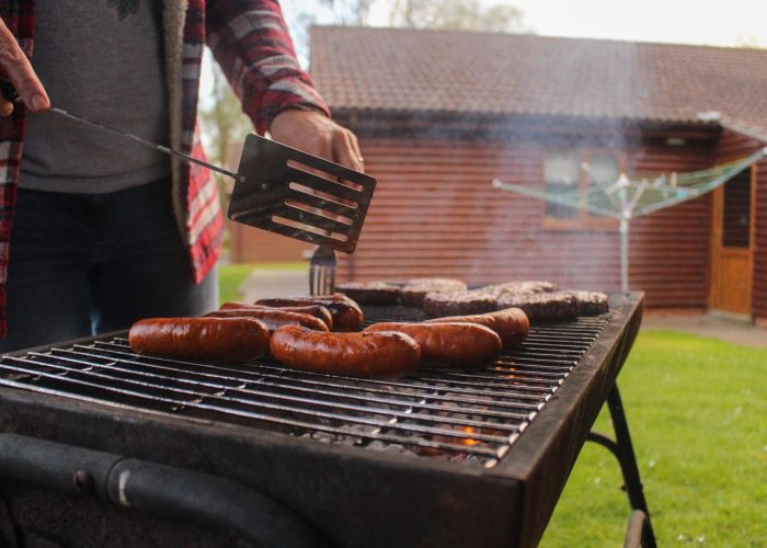Grilling sausages and patties in a backyard with a rustic wooden house in the background.